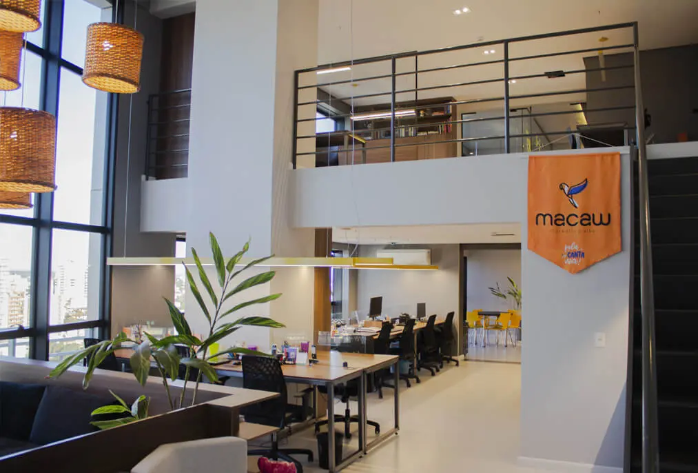 O escritório da Macaw, iluminado por grandes janelas, com mesas largas e uma escada que leva a um mezanino.