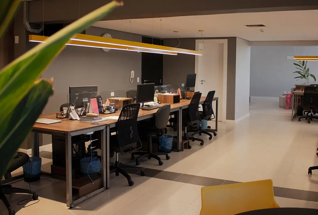 Visão de trás de uma planta alta do escritório da Macaw: mesas largas com cadeiras de escritório e monitores desligados. Ao fundo, uma porta para um banheiro e o arco de entrada que leva às poltronas.