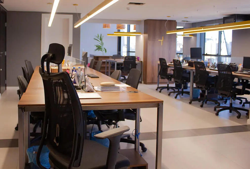 Agência da Macaw vista do ponto mais ao fundo do escritório: mesas largas com cadeiras e material de escritório, iluminadas por lâmpadas de LED compridas.